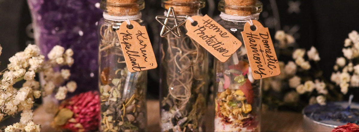 Three small glass vials with cork stoppers containing dried botanicals and a rolled letter, each with a handwritten paper tag reading names like 'Karen Repellant', 'Home Protection', and 'Self Love', styled on a wooden surface with dried flowers and crystals.